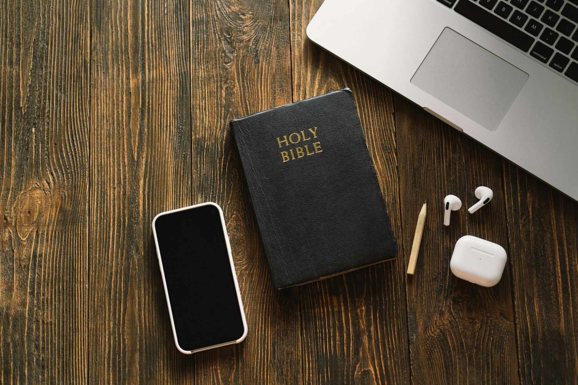 Holy Bible, smartphone, laptop, earbuds, and pencil on wooden desk, symbolizing tech and spirituality connection.