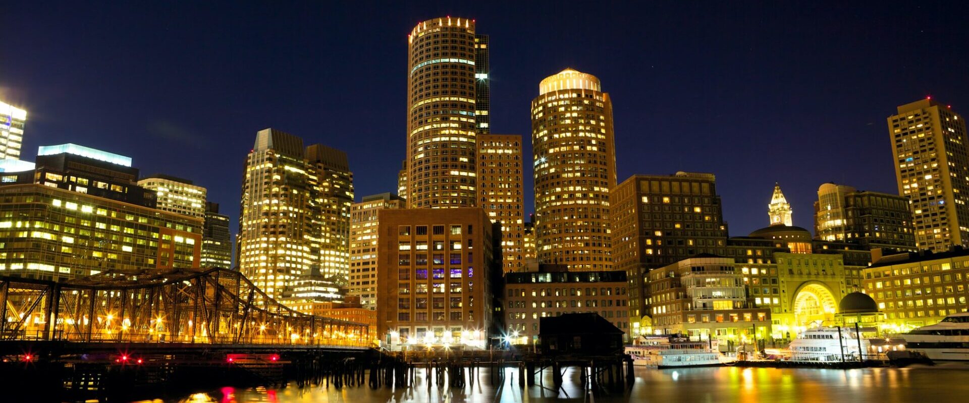 Boston skyline at night, glowing skyscrapers reflecting on the harbor, highlighting the city's vibrant urban atmosphere.