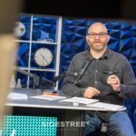 Man speaking into microphone in a studio with a modern blue geometric wall background.