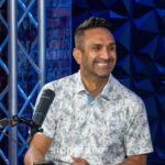 Smiling man in patterned shirt, sitting with microphone in a blue-themed studio setting.