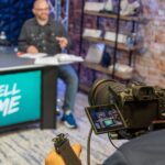 Professional video shoot setup with host at desk, camera in the foreground, and studio shelves in the background.