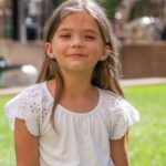 Smiling girl in a white blouse enjoys sunny day in a park, with green background and natural light.