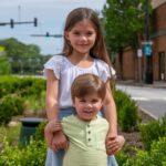 Two smiling kids posing outdoors on a sunny day with greenery and buildings in the background.