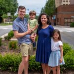 Happy family of four posing outdoors on a sunny day, with buildings and greenery in the background.