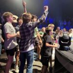 A group of young people enthusiastically cheering at a vibrant indoor event under colorful lighting.