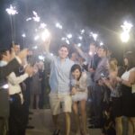 Wedding couple walking through sparkler-lit path surrounded by cheering guests at night celebration.