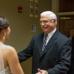 Father and bride share a joyful moment before the wedding ceremony, with family in the background.