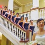 Bride and bridesmaids on staircase, wearing black dresses and white gown, smiling at a wedding venue.