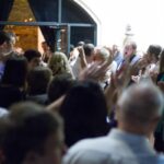 Celebratory crowd at an evening event, people clapping and cheering under warm lights in a festive atmosphere.