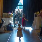 Little girls walking towards wedding guests, gifts table in background, indoors with bright doorway.