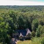 Aerial view of house nestled in lush green forest, surrounded by dense trees under bright blue sky.