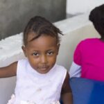 Young girl smiling softly, wearing a light dress, sitting by a blue table with another child in a bright pink shirt.