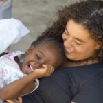 A woman joyfully holds and plays with a laughing child while seated outdoors, capturing a moment of happiness.