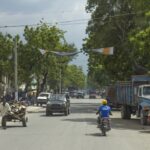 Busy street with motorcycles and trucks under green trees on a sunny day.