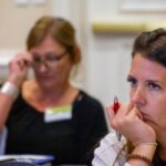 Woman attentively listening in a conference setting, holding a pen, with another person blurred in the background.