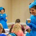 Women in vintage blue dresses enjoying coffee and laughter at a social event indoors.