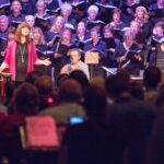 Choir performing on stage with soloist in front, wearing red, singing passionately to a large audience.