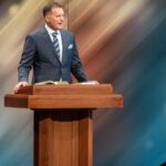 Man delivering a speech at a podium, wearing a suit and tie, with a blurred background.