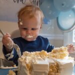 Baby joyfully smashing cake at first birthday party, surrounded by blue balloons and decorations.