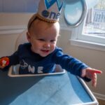 Smiling baby with a one crown celebrating first birthday in high chair with blue and white decorations.